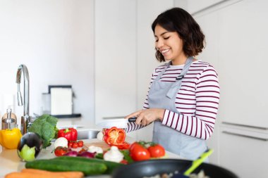 Happy beautiful young brunette middle eastern lady wearing grey apron enjoying cooking at home, woman cutting vegetables and smiling, preparing healthy meal, using only organic veggies, copy space