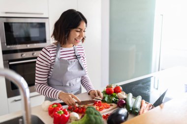 Cheerful pretty brunette young hispanic woman enjoying cooking at home, reading recipe, food blog on Internet, using modern digital tablet while preparing healthy meal at kitchen, copy space