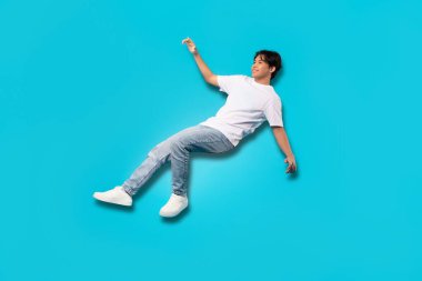 Happy Asian Teenager Boy Falling Posing In Mid Air Smiling Looking Aside Over Blue Background. Studio Shot Of Positive Teen Guy Experiencing Zero Gravity. Full Length