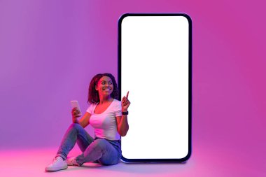 Smiling Black Woman With Smartphone In Hand Sitting In Neon Light And Pointing At Huge Blank Telephone Screen, Happy African Female Recommending New Mobile Application, Creative Collage, Mockup