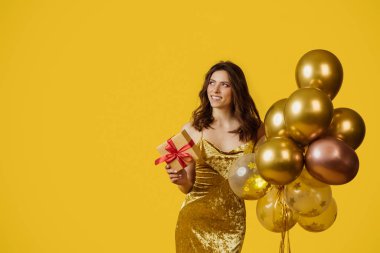 Birthday celebration concept. Charming young lady in festive dress with balloons and gift box looking aside and smiling over yellow studio background, copy space