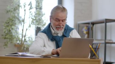 Positive retirement. Cheerful senior grey haired man in eyeglasses web surfing online on laptop computer, enjoying great news, sitting at home, tracking shot, slow motion, free space