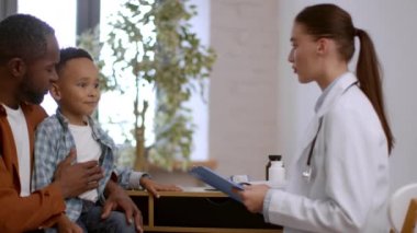 Kids healthcare. Lovely african american boy sitting at doctor office with daddy, woman pediatrician measuring patient body temperature and writing in medical history, tracking shot, slow motion