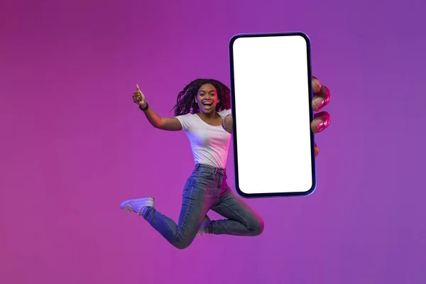 Excited Black Woman Jumping With Big Blank Smartphone In Hand And Showing Thumb Up, Happy African Lady Recommending New App Or Online Offer While Having Fun Under Neon Light, Collage, Mockup