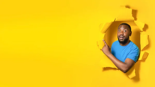 Shocked black man pointing finger aside at free space for text or design, posing in hole in torn of yellow paper background. Look there concept. Panorama
