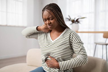 Unhappy young black woman wearing casual clothes suffering from menstrual cramps, feeling sick to her stomach, holding belly and touching neck, pain during period, sitting on couch at home