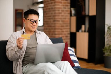 Happy middle aged asian man using credit card for online shopping, sitting on sofa with laptop, home interior, free space. Excited male paying for goods and services online