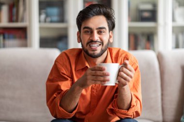 Cheerful handsome middle eastern millennial man in smart casual sitting on couch in living room, enjoying fresh morning coffee at home, smiling at camera, copy space. Domestic lifestyle concept
