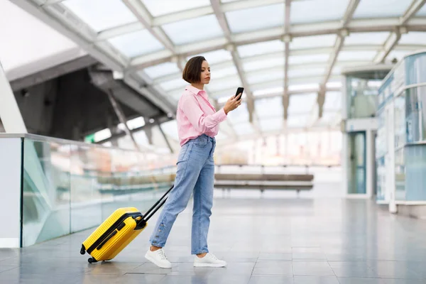 Beautiful Young Woman With Suitcase Walking At Airport Terminal And Using Smartphone, Female Traveller Texting On Mobile Phone Or Browsing Internet While Going To Departure Gate, Copy Space