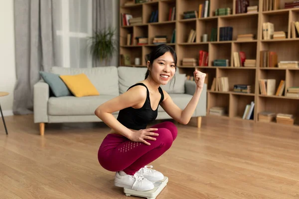 Sports and healthy diet for weight loss concept. Young korean lady in sportswear sitting on scales and making YES gesture, happy with result of her slimming diet