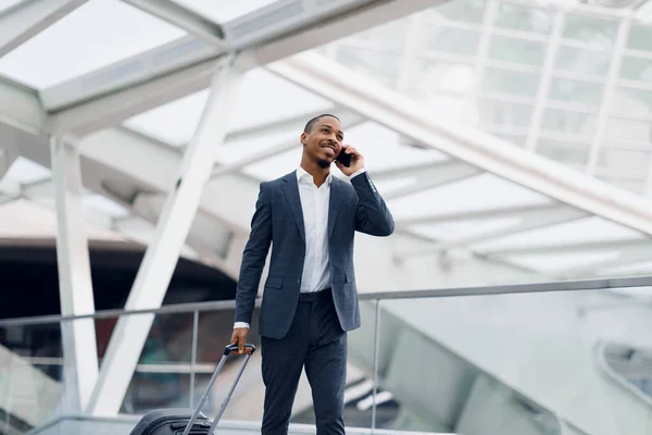 Smiling Black Businessman Talking On Cellphone While Walking With Suitcase In Airport, Handsome African American Male Enjoying Mobile Phone Conversation While Going To Flight Gate, Copy Space