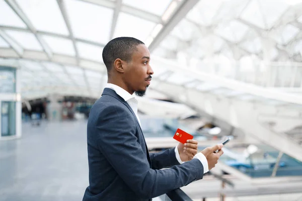 Young Black Man Relaxing With Smartphone And Credit Card While Waiting Flight At Airport, Millennial African American MaleHolding Mobile Phone And Looking Away While Standing At Terminal Hall