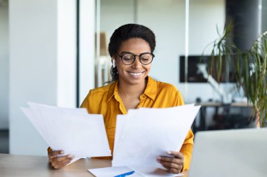 Excited black businesswoman reading papers and positive business report, sitting at workplace in modern office interior. Successful entrepreneurship career, accounting and documentation