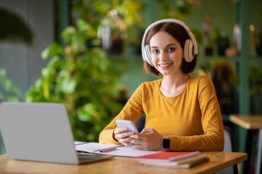 Pretty young smiling woman using wireless headphones and phone, listen to music while work or study on laptop pc computer, sit at table in coffee shop cafe indoor. Freelance business concept