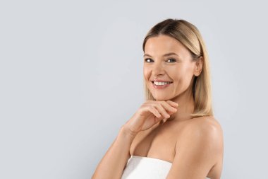 Positive cheerful middle aged pretty blonde lady wrapped in white towel posing on grey studio background, woman touching her smooth glowing face skin, copy space. Cosmetology concept
