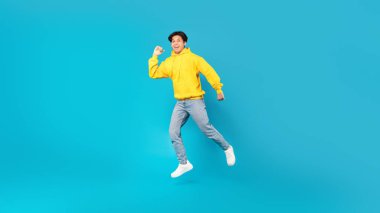 Joyful Chinese Teen Boy Jumping Smiling To Camera Posing Over Blue Background In Studio. Teenager Guy Celebrating Success Leaping In Mid Air Wearing Casual Clothes. Panorama, Full Length Shot