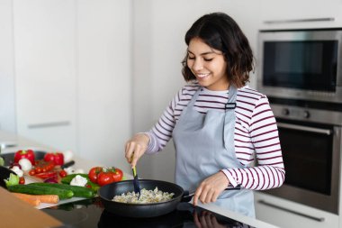 Beautiful young middle eastern woman holding cooking spatula, mixing rice with vegetables in frying pan on electric stove, preparing food at kitchen, copy space. Healthy diet, nutrition