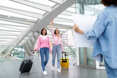 Two females walking with suitcases and waving to friend waiting for them with name board at airport after arrival, happy women smiling, feeling excited for long-awaited meeting, selective focus