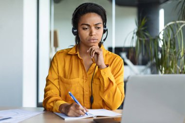 Online business education. Focused black woman in headset studying with laptop in office, watching webinar and taking notes to notepad, enjoying distance learning