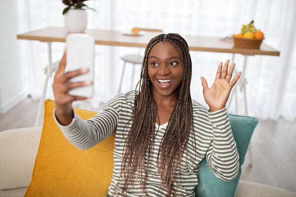 Cheerful happy attractive young long-haired black lady in homewear sitting on couch in living room at home, talking with girlfriend online, using smartphone, copy space. Telecommunication concept