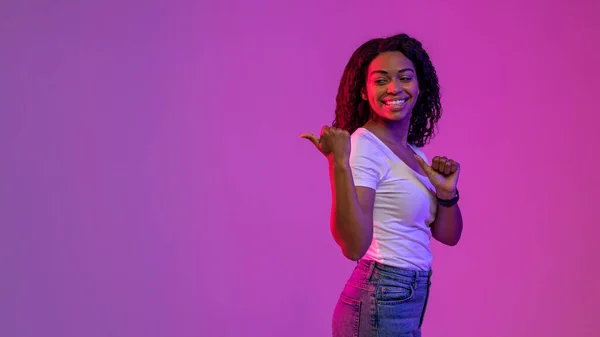 Great Offer. Smiling Black Woman Pointing At Copy Space Behind Her Back With Two Thumbs Up, Happy African Female Showing Free Place While Standing In Neon Light Over Purple Background, Panorama