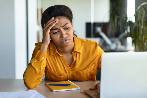 Unhappy african american female manager feeling sad, sitting at workplace and looking at laptop in office interior. Business and problems at work, mistake, overworked