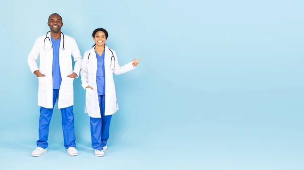 Excited black male and female doctors posing over blue background, woman pointing aside at copy space, advertising medical advertisement, standing over blue background. Healthcare and medicine