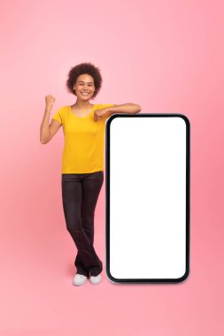 App and website advice. Glad young african american curly lady show victory and success gesture near huge smartphone with empty screen, ad and offer isolated on pink background, studio. Blog and chat