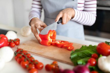 Female hands cutting fresh organic red bell pepper, unrecognizable woman wearing apron housewife preparing healthy meal, chopping fresh vegetables, kitchen interior, cropped