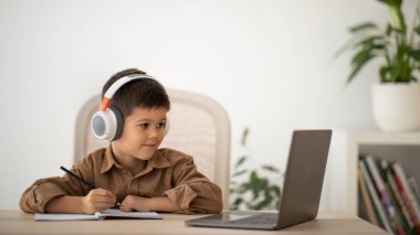 Cheerful european small kid in headphones has video call with teacher, tutor at table in living room, elementary school and kindergarten interior. Device for modern education for children at home