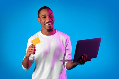 Happy bearded handsome young african american man in white holding modern laptop and credit card over neon studio background, smiling at camera, shopping on Internet, purchasing goods, copy space clipart