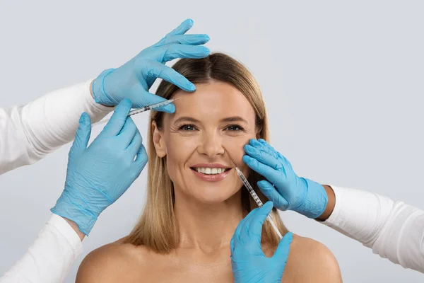 Closeup of smiling blonde beautiful middle aged woman get beauty injections in her face, doctors hands in medical gloves holding needle next to adult female patient face, studio background