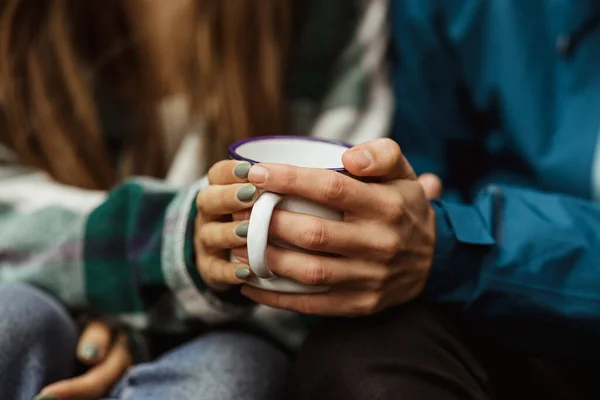 Hands of millennial caucasian woman and guy in jackets hold cup of hot drink, travel in cold forest, enjoy adventure together outdoor. Rest, vacation, active lifestyle and camping, hikers and walk