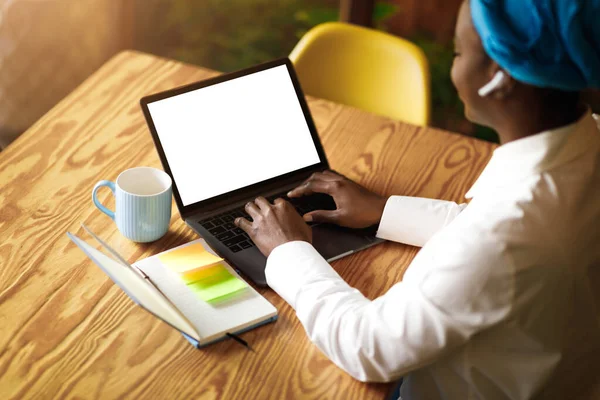 Remote job concept. Unrecognizable black woman in smart casual freelancer typing on laptop with white blank screen, using earpods, businesswoman working at cafe, high angle shot, mockup