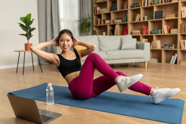 Young japanese lady exercising at home, doing abs exercises, working out with laptop computer. Fit korean woman in sportswear strengthening her core muscles
