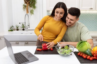 Handsome young brunette guy helping his pretty girlfriend making dinner at home, happy beautiful couple cooking together at modern cozy kitchen, using laptop, reading food blog, copy space