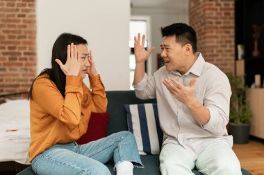 Marital problems, divorce concept. Married japanese couple having fight, yelling at each other, sitting on sofa in living room. Furious man and woman shouting at one another