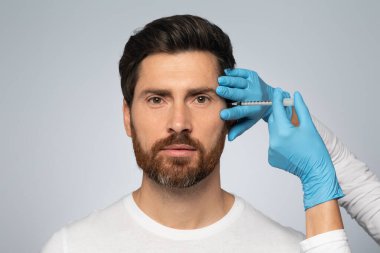 Handsome bearded man getting under eye injections in beauty clinic, looking at camera, standing on grey studio background. Male attending beautician, having face filler