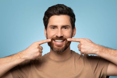 Handsome bearded man in t-shirt posing over blue background, smiling and pointing with fingers at perfect teeth. Dental health concept