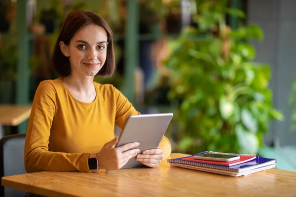 Remote job, freelance concept. Pretty cheerful young brunette woman with nice haircut sitting at table, smiling at camera, holding modern digital pad, working at coffee shop, copy space