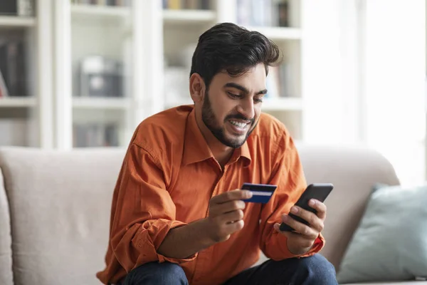 E-commerce, retail, financial app. Happy handsome young arab guy in smart casual banking or shopping from home, sitting on couch in living room, using smartphone and credit card, copy space