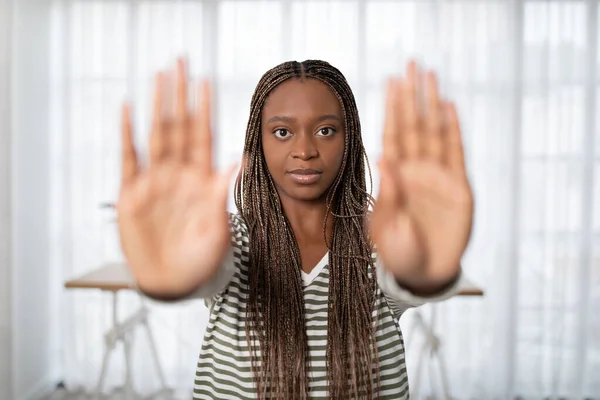 Concerned beautiful young african american long-haired woman in casual outfit showing both palms stop or no gesture at camera, home interior. Stop domestic abuse, violence concept