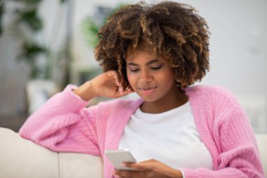 Closeup of positive cheerful relaxed pretty curly millennial black woman sitting on couch at home, using modern smartphone, chatting with guys on dating mobile app, copy space