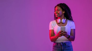 Smiling Black Female Holding Smartphone And Looking Aside At Copy Space While Standing In neon Light Over Purple Background, Happy African American Lady Enjoying Mobile Offer, Panorama