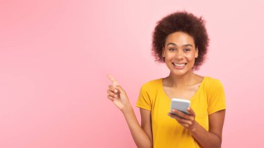 Laughing shocked young black curly lady with smartphone pointing her finger at empty space isolated on pink background, studio, panorama. Idea creation, app, blog, recommend sale online, ad and offer