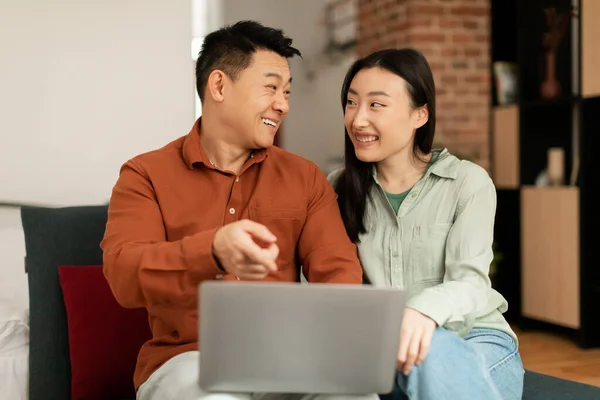 Digital entertainment. Excited asian couple using laptop at home, smiling middle aged man and young woman relaxing in living room with computer, watching movies, male pointing at screen