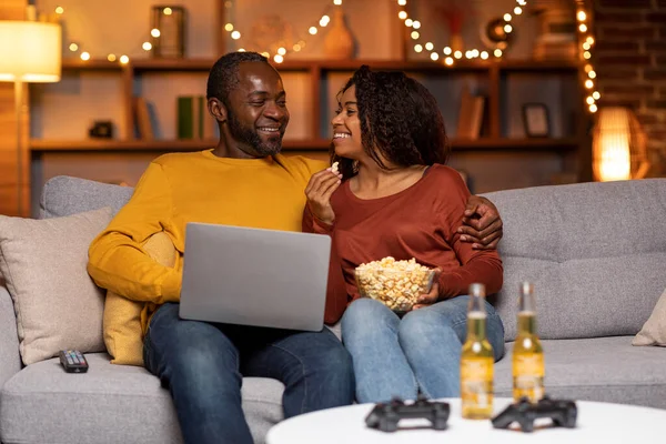 Happily married cheerful african american couple sitting on couch, using laptop at cozy winter evening, watching movie online, eating popcorn, drinking beer, resting at home together, free space