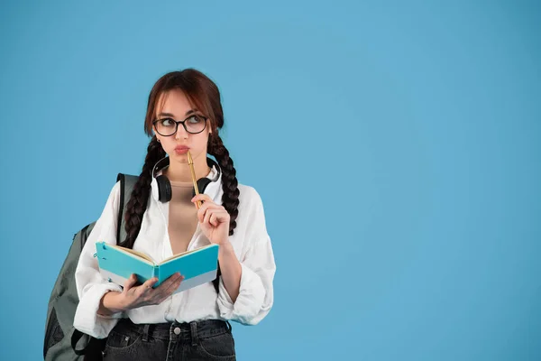 Pensive smart inspired caucasian teen girl with pigtails in glasses with headphones student with backpack holding notebook, thinks, isolated on blue background, studio. Learn, knowledge, education