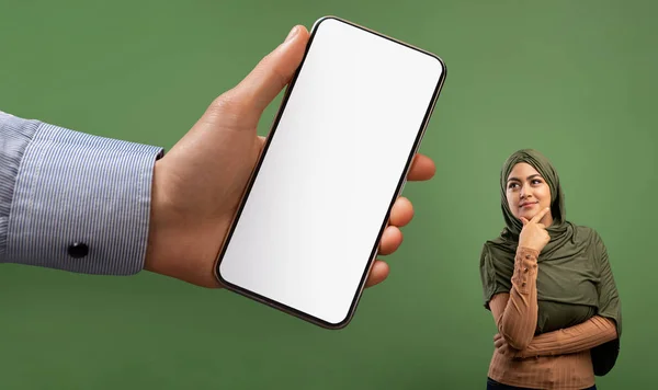 Thoughtful Muslim Woman Looking Aside At Blank Smartphone In Huge Male Hand, Giant Arm Holding Cellphone With White Screen And Showing To Curious Islamic Lady In Hijab Over Green Background, Mockup