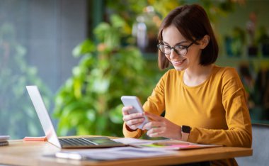 Pretty smiling young woman in smart casual and eyeglasses sitting at table, using modern laptop and smartphone at cafe, female entrepreneur using business mobile app, copy space